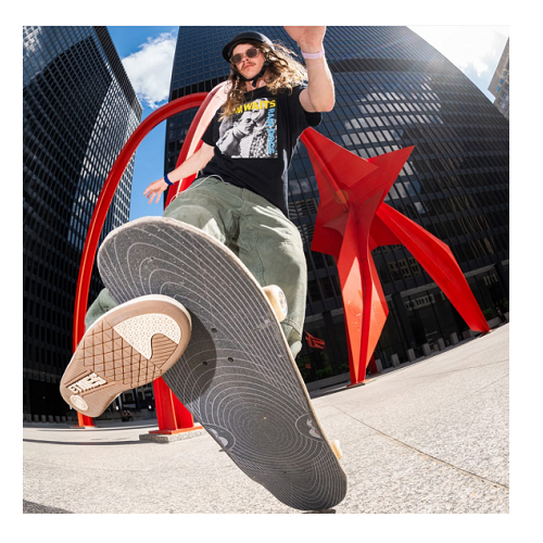 A photo of Andy Anderson doing a skateboard trick in front of a glass skyscraper building and red sculpture behind him. 